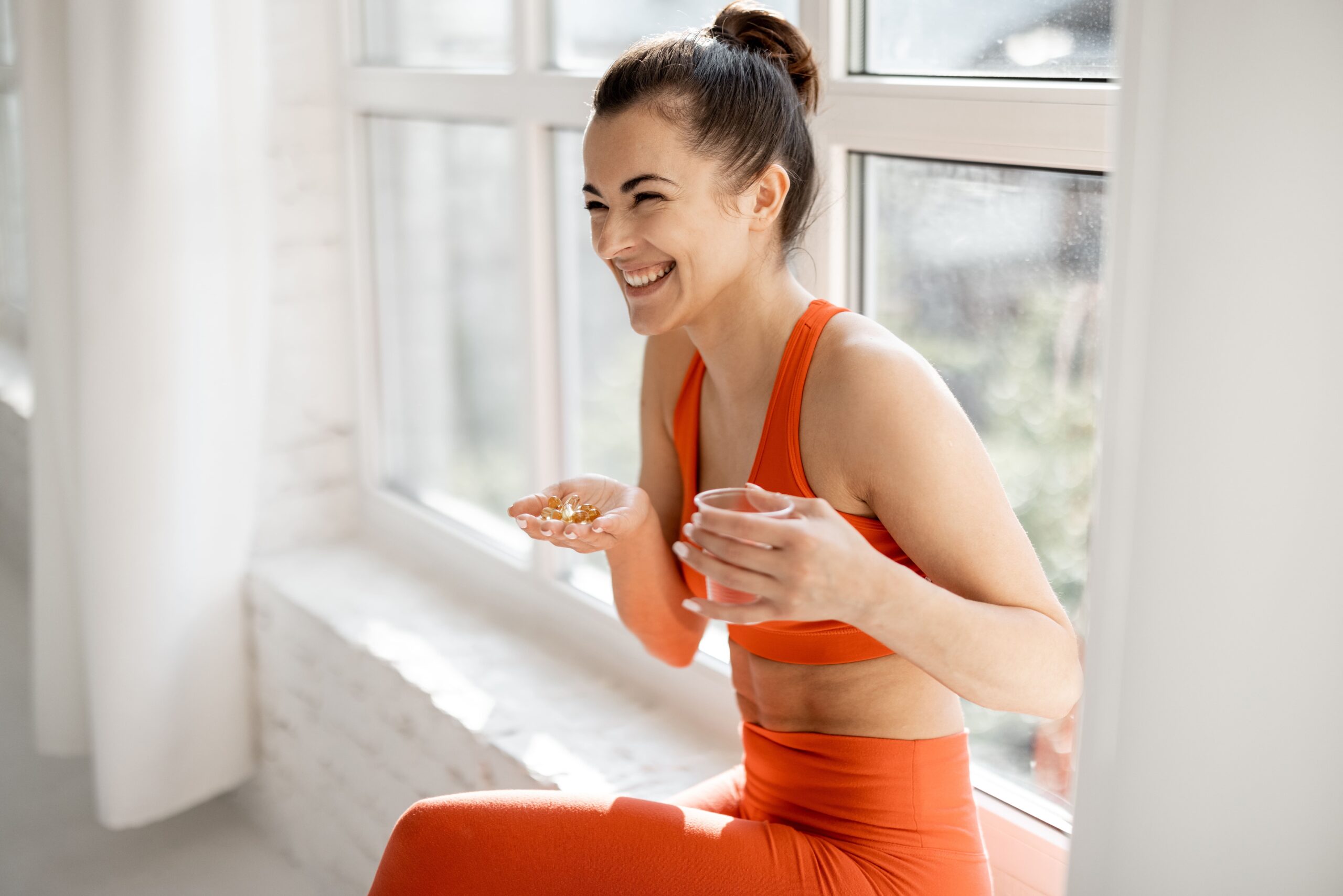 Mulher com roupa de academia na cor laranjada com um copo de água em uma mão e na outra várias capsulas de suplementos vitamínicos.