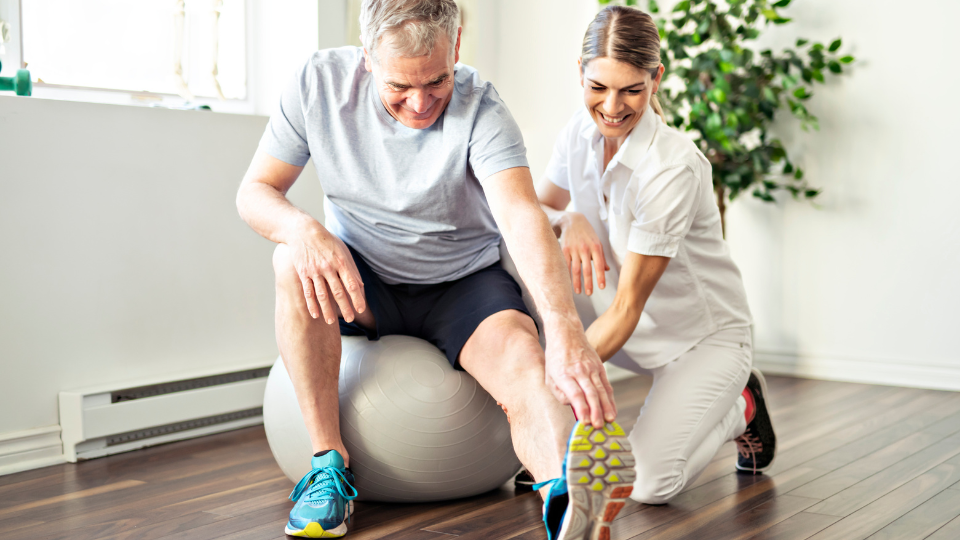 Homem e mulher com vestimenta de profissional de saúde. O homem está com roupa de atividade física e tênis, fazendo alongamento segurando na ponta do pé.