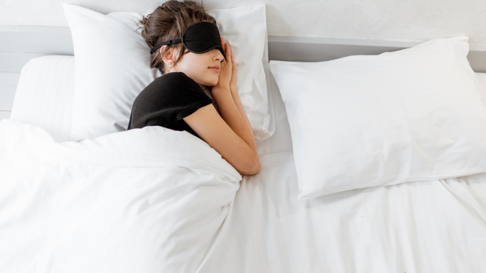 Mulher dormindo em uma cama com lençóis e travesseiros brancos, usando blusa preta e tapa olho.