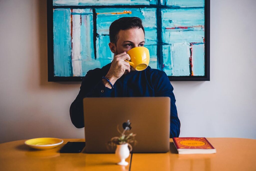 Homem tomando café em uma xícara amarela em frente ao computador aproveitando benefícios da cafeína